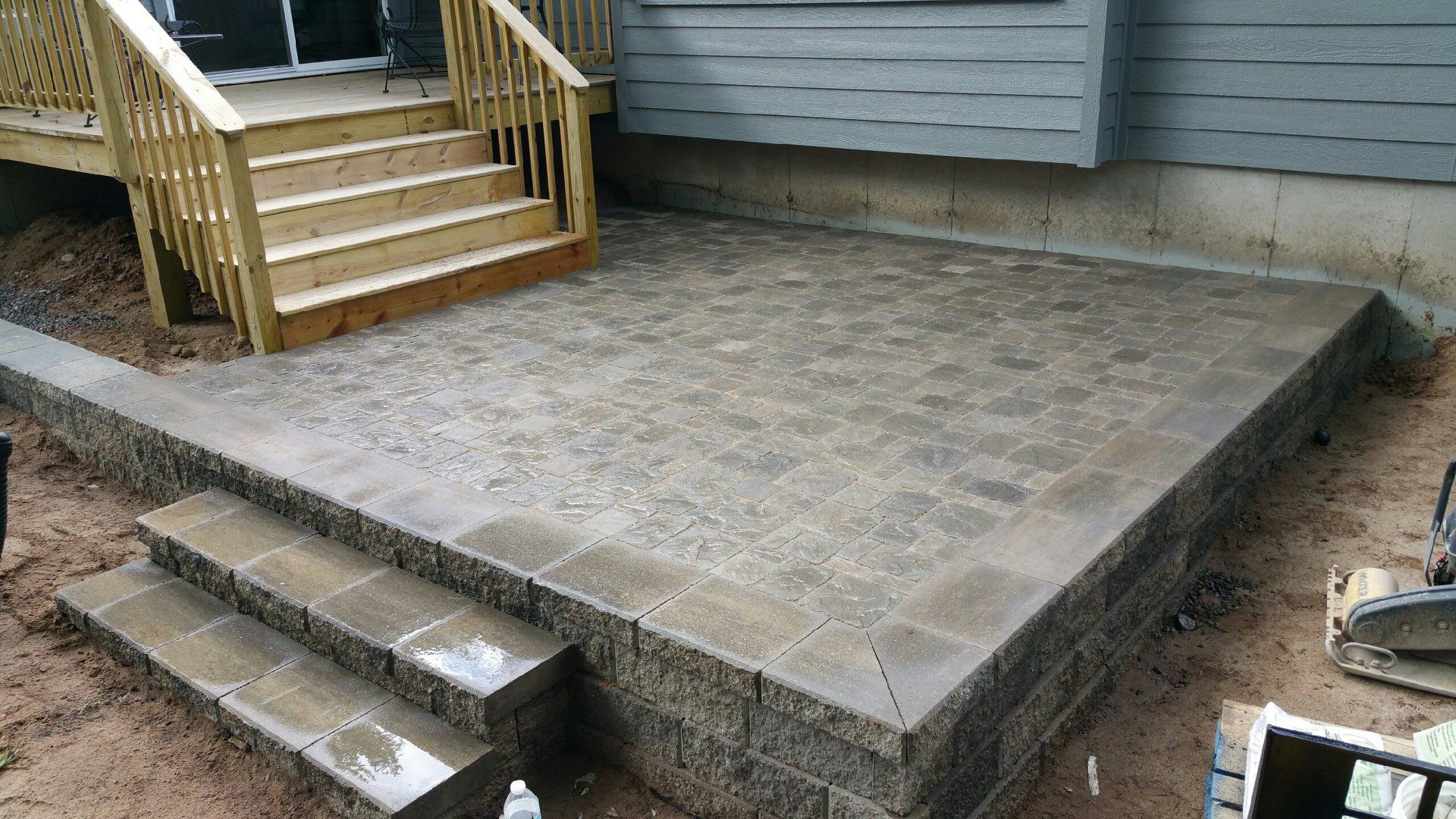 Stone patio with retaining wall and wooden stairs leading up to a deck and house.