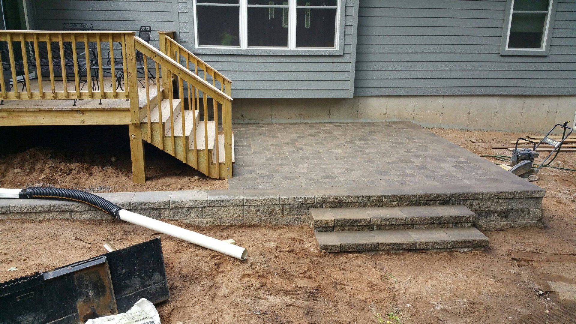 Deck with steps, a stone patio, and a drain pipe next to a gray house.