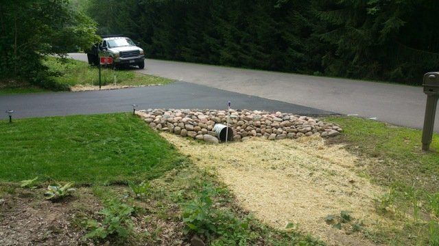 Rock-lined drainage area with a metal pipe under a driveway. A truck is parked on the road.
