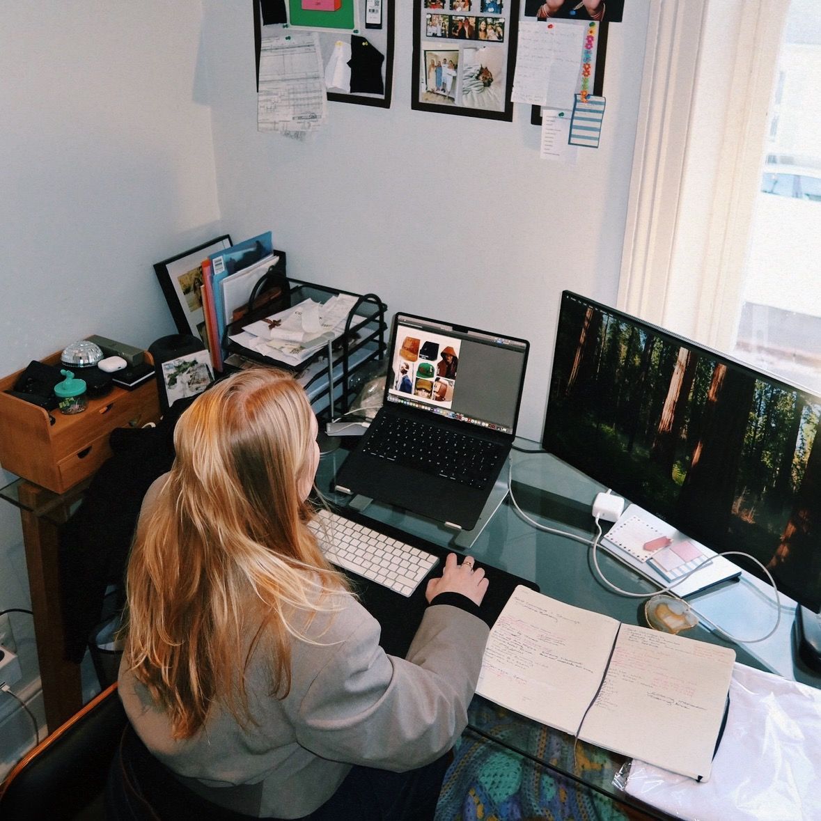 Woman at desk with laptop, monitor, and notebook; working.