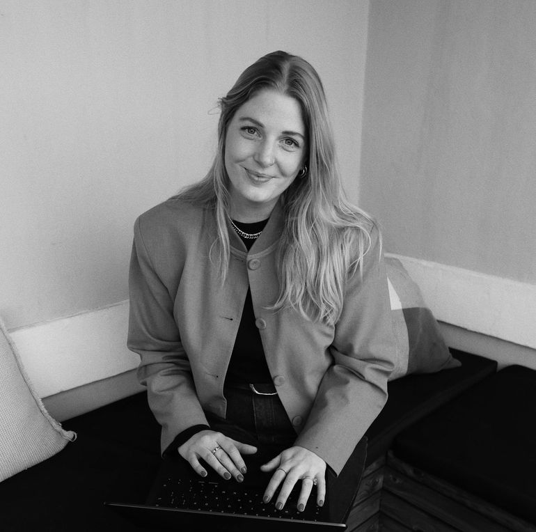 Woman in blazer seated with laptop, smiling. Inside, couch setting.