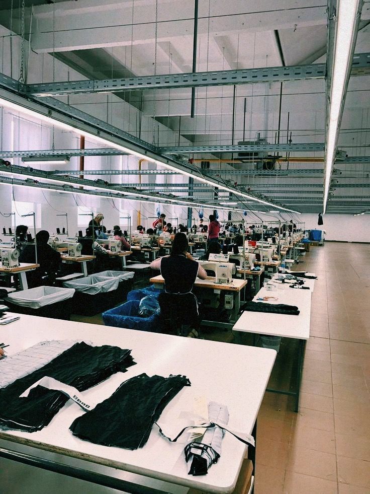 Large sewing factory with workers at tables, creating clothing. Fluorescent lights illuminate the space.