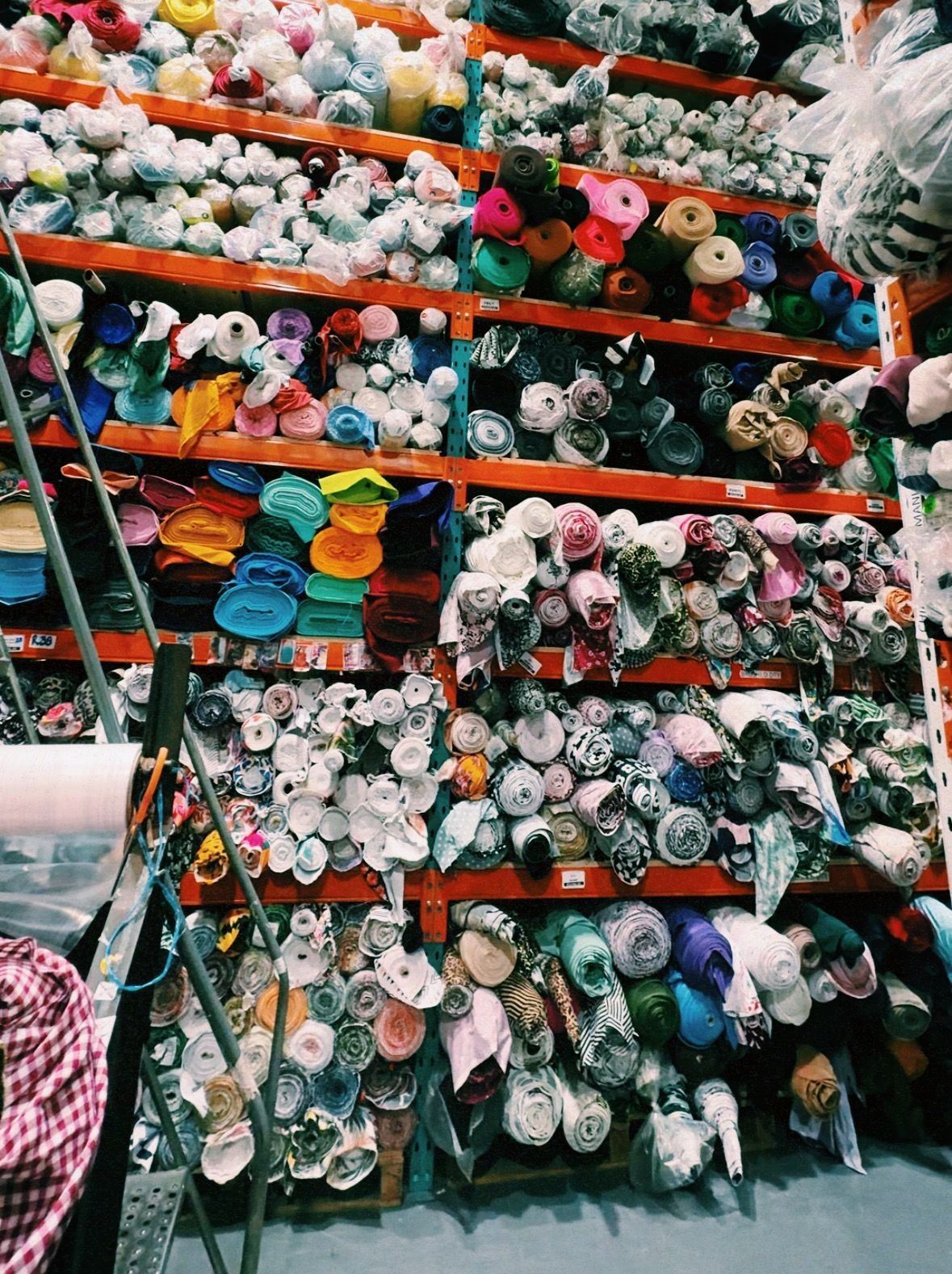 Fabric rolls stacked on shelves in a warehouse; many colors and patterns. Ladder leans against the shelves.