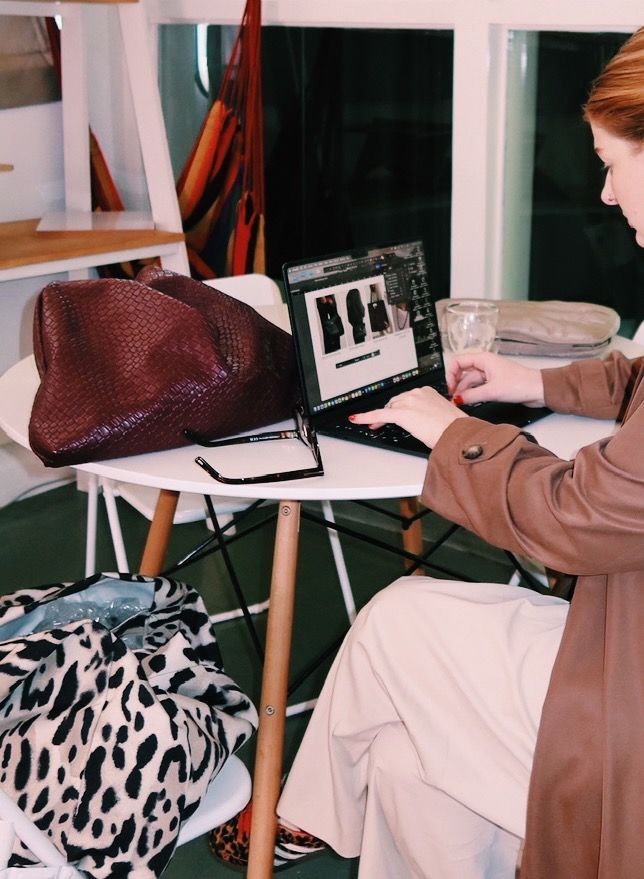 Woman working on a laptop at a white table. A large purse and leopard-print bag are nearby.