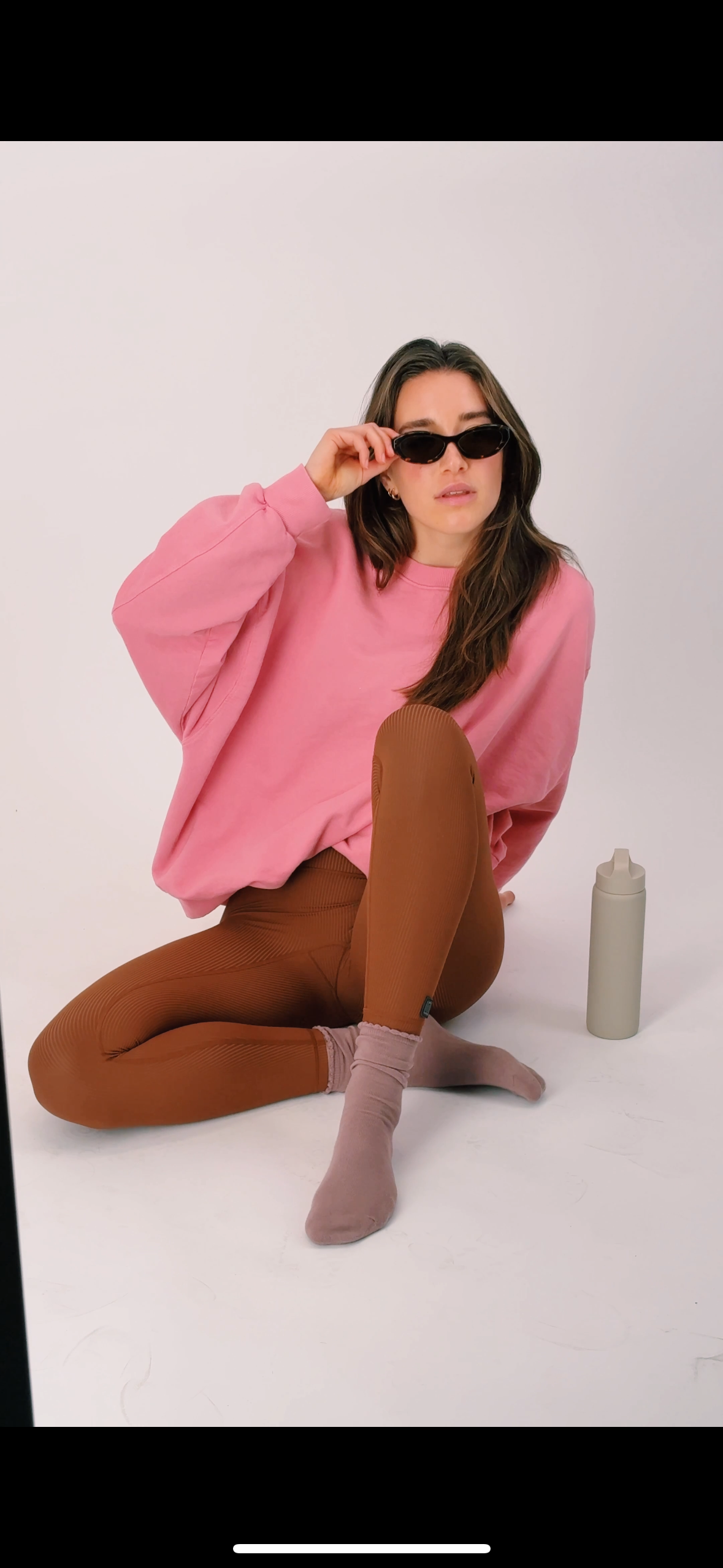 Woman in pink sweatshirt and brown leggings, wearing sunglasses, seated.