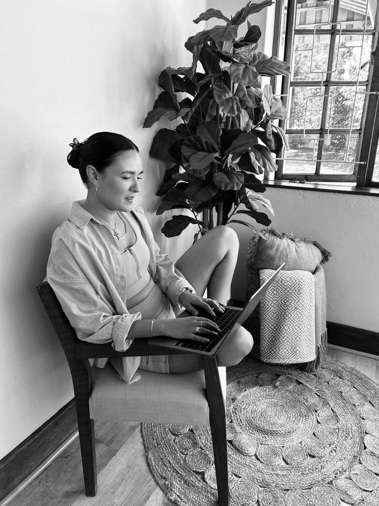 Woman sitting in chair, working on laptop near window and plant.