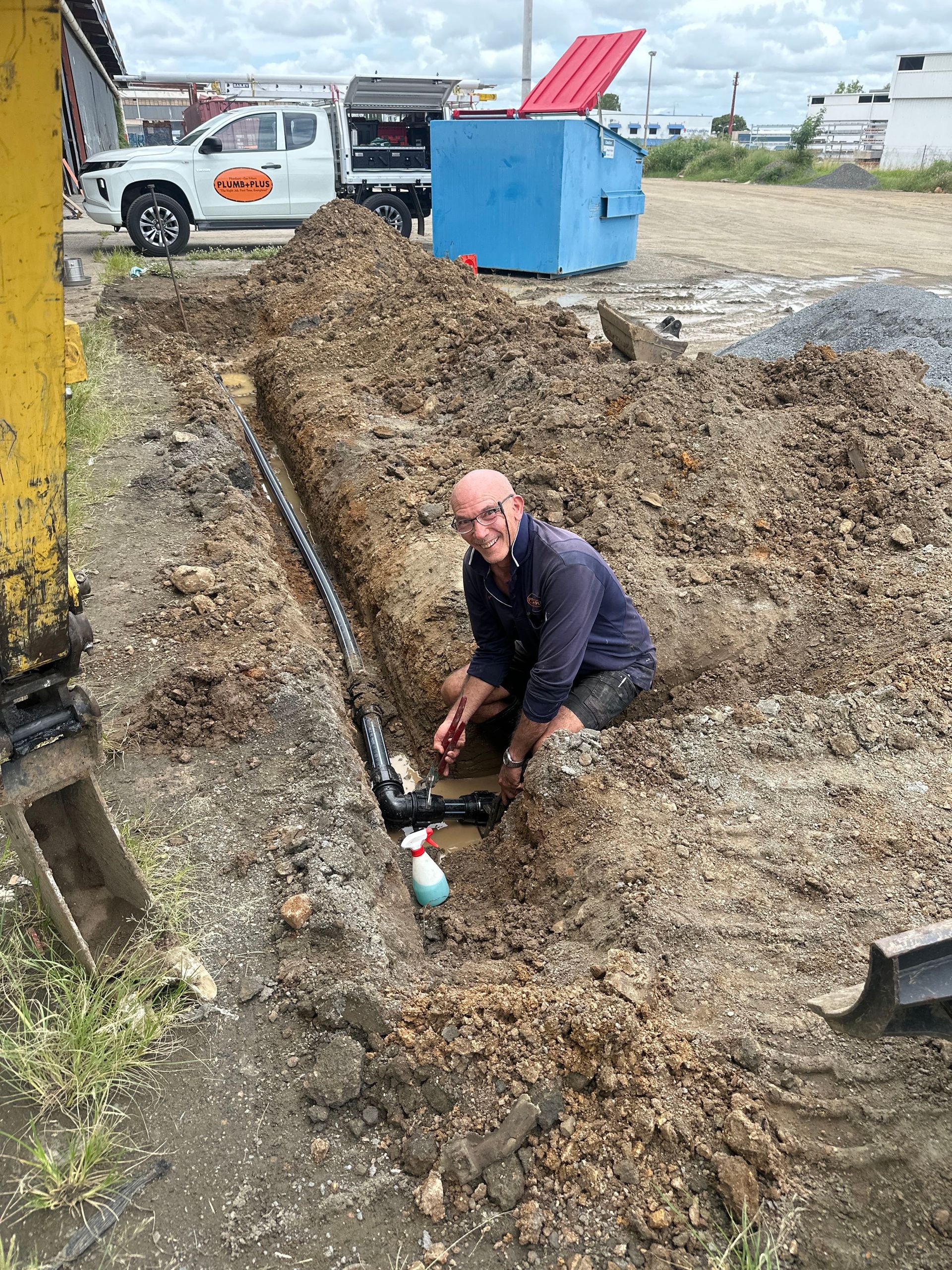Worker crouches in a muddy trench near a blue utility box and parked truck.
