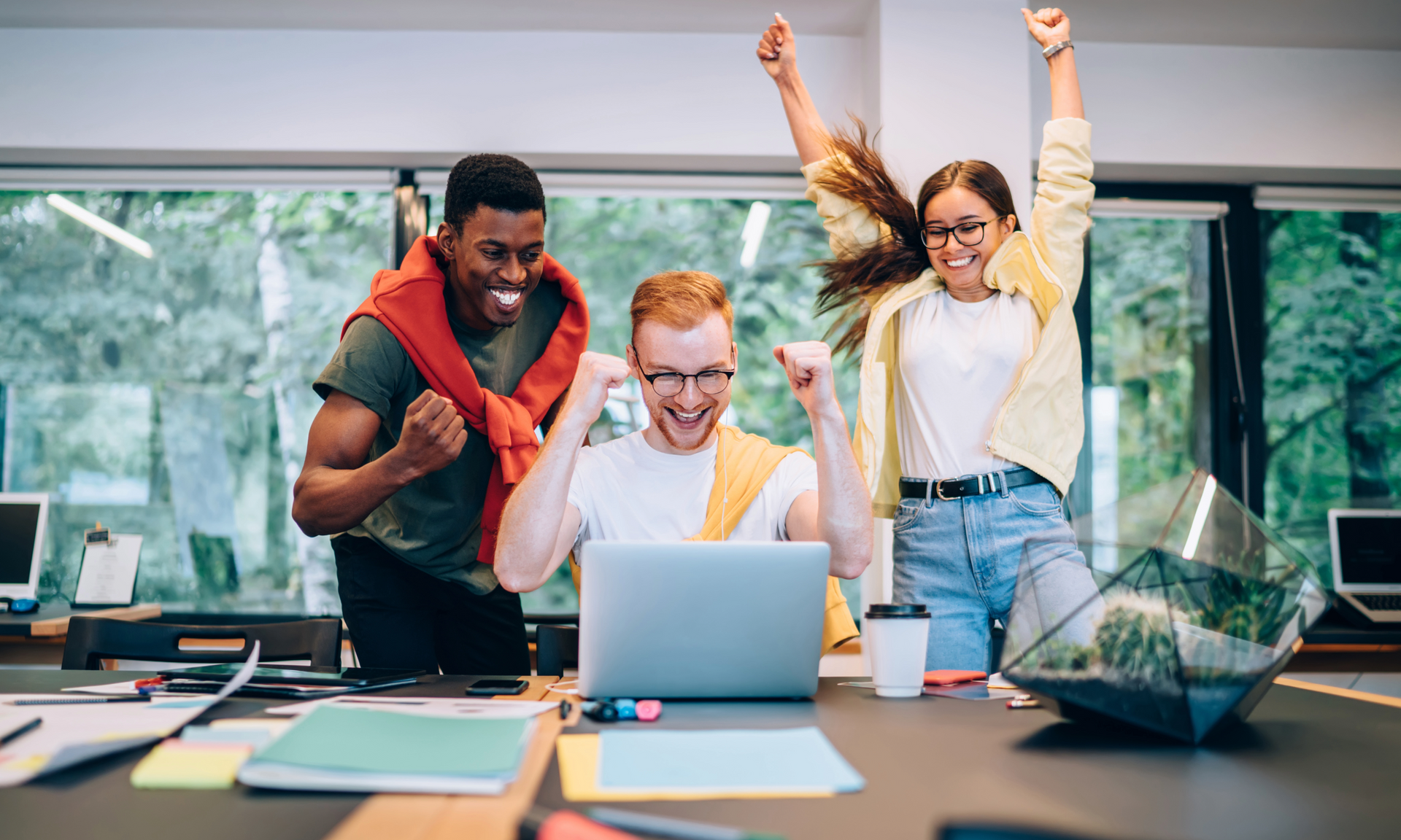 Drei junge Menschen jubeln begeistert vor einem Laptop in einem modernen Büro, ihre Freude deutet auf einen gemeinsamen Erfolg oder Durchbruch hin.