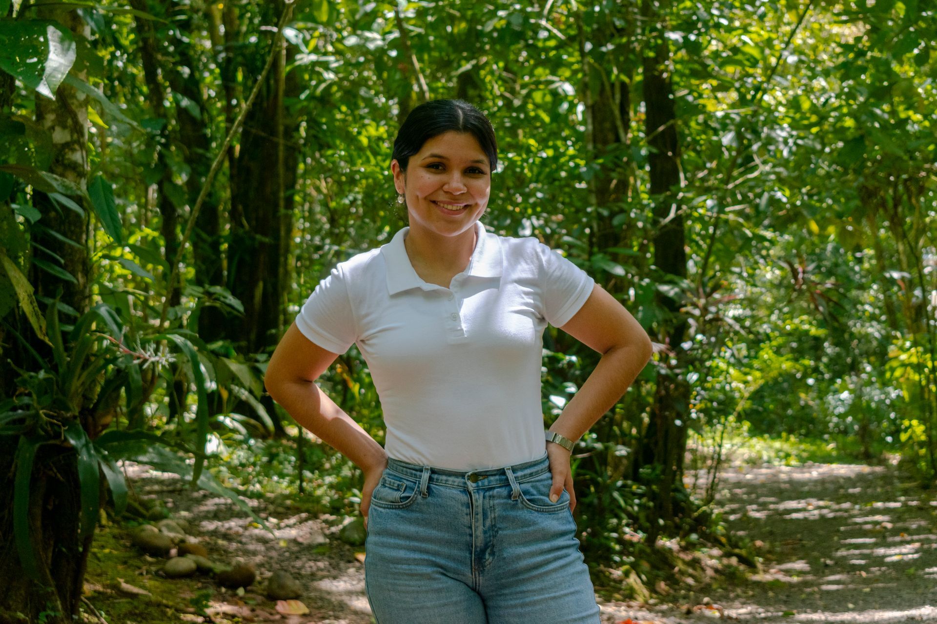 Woman in white shirt and jeans poses in a lush, green forest setting, hands on hips.