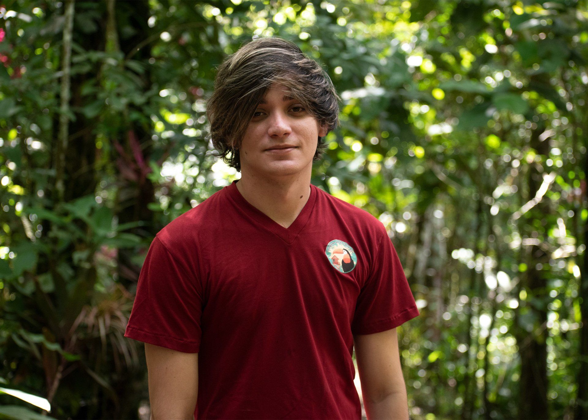 Young man with dark hair in a red shirt smiles in a green, leafy outdoor setting.