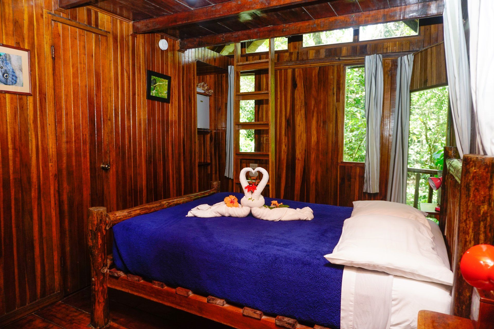 Wooden bedroom with a blue bedspread, towels shaped as swans, and a window overlooking green foliage.