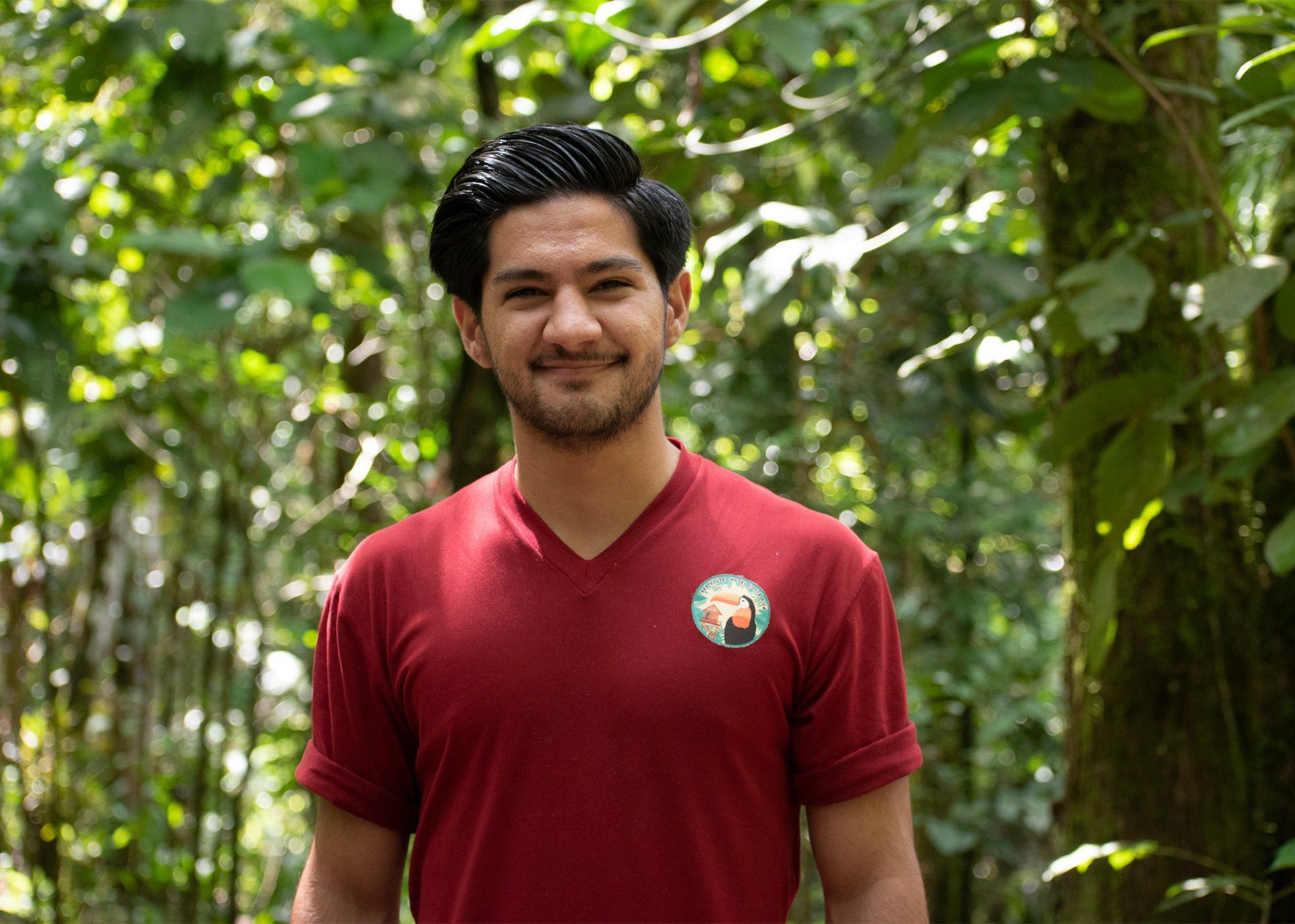 Man in red shirt smiles in a lush green forest.