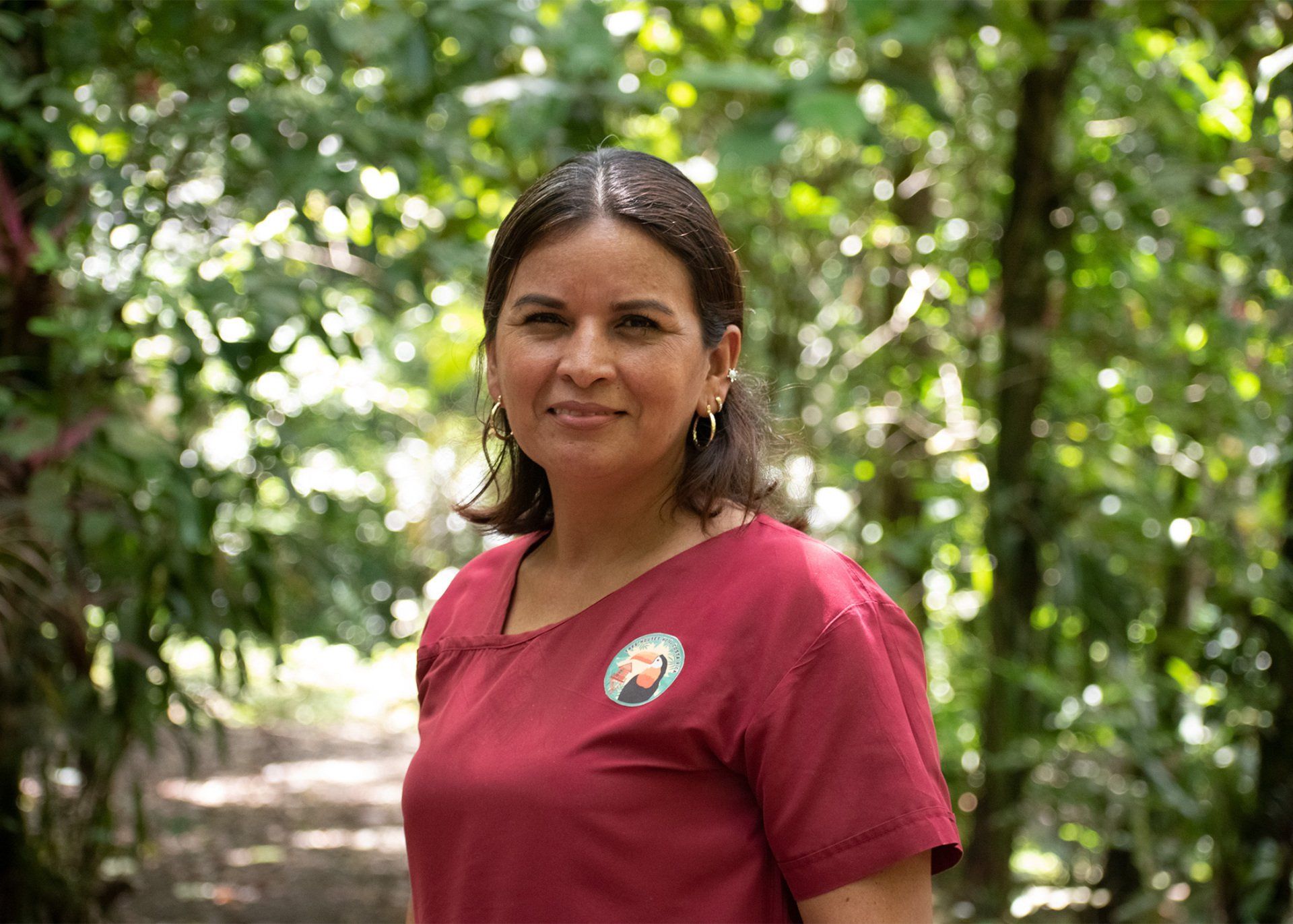 Woman in maroon shirt smiles in a forest, with green foliage behind her.