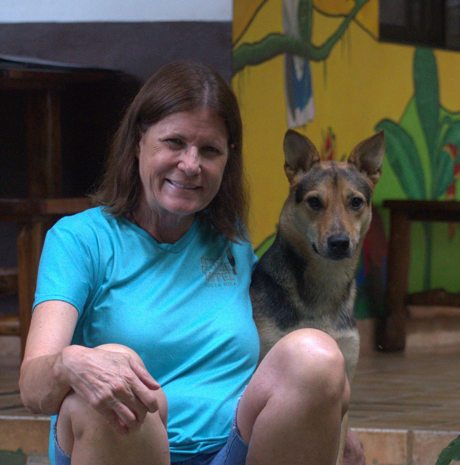 Woman in blue shirt sitting with a dog, both looking forward; outdoors with colorful mural.