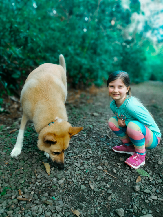 Girl squats smiling next to dog sniffing ground on a gravel path in a wooded area.