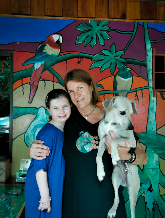 Woman and girl pose with dog in front of colorful mural of birds and foliage.