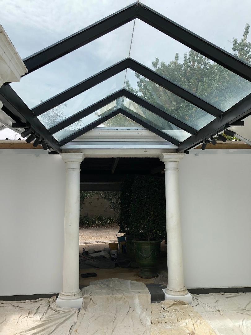 White courtyard with a black glass-roofed pergola and central doorway framing a shaded outdoor space