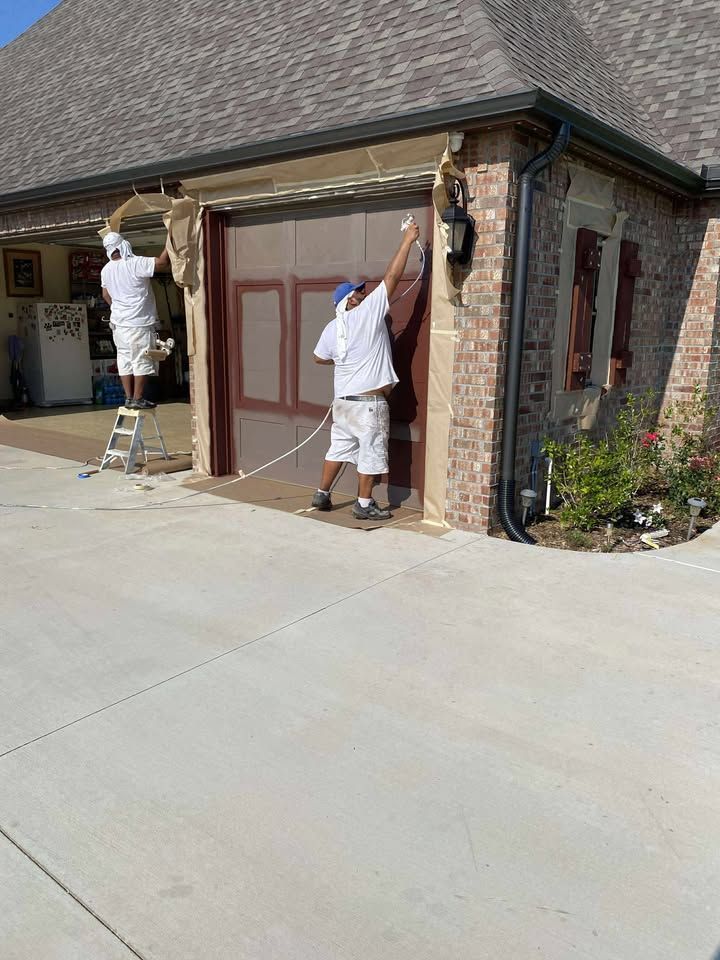 Two painters in white uniforms repainting a garage exterior beside a brick house