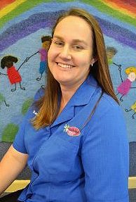 A woman in a blue shirt is smiling in front of a rainbow rug