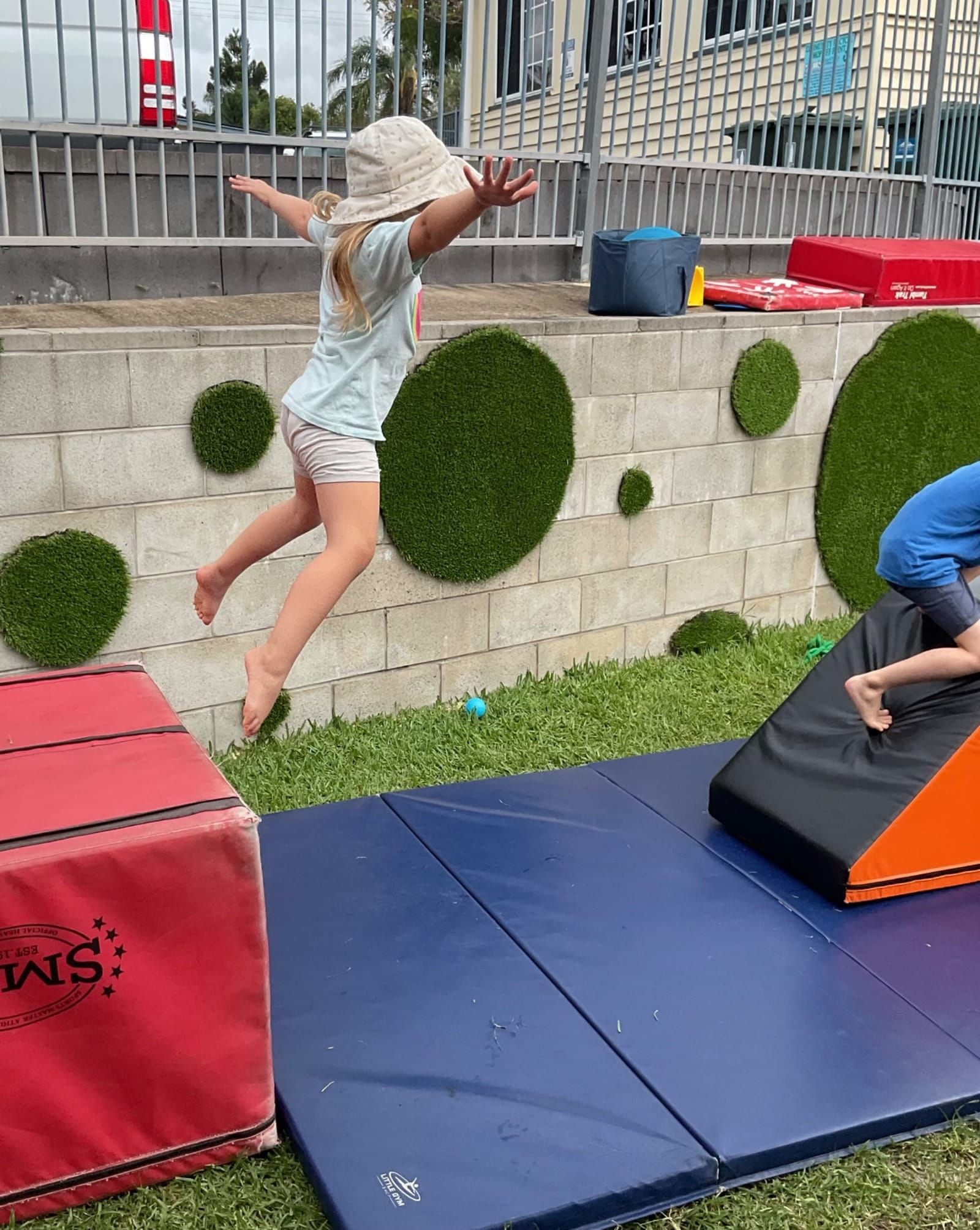 A girl is jumping in the air while a boy is sitting on a mat.