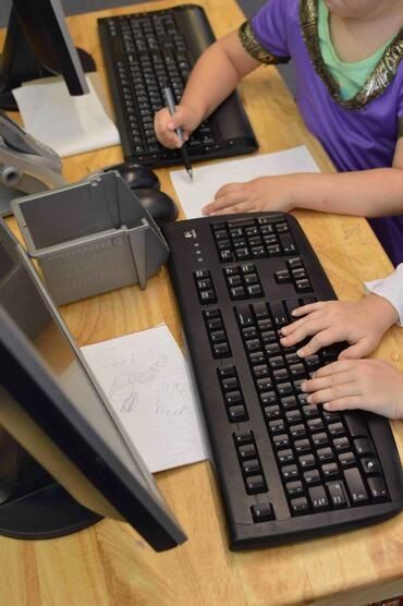 Two children are typing on a keyboard in front of a computer monitor