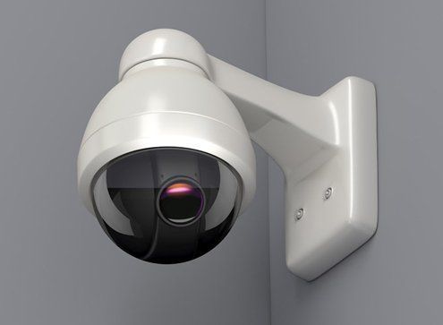 White security camera mounted on a gray wall, in a corner, with a dark lens reflecting red light.