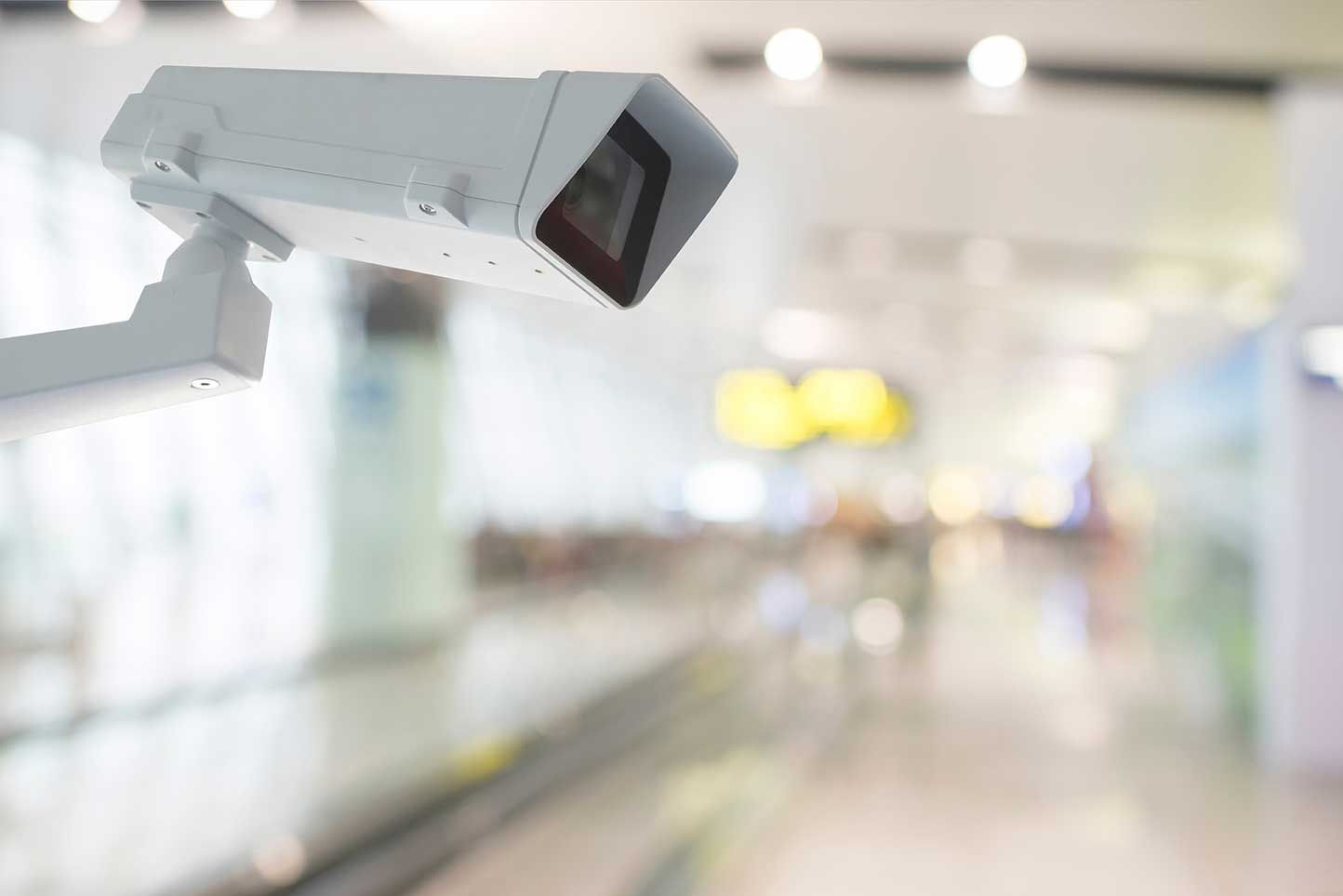 White security camera mounted in an airport, with a blurred background of a walkway and bright lights.