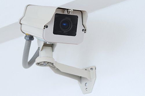 Security camera, cream-colored, mounted on a white wall, angled toward the viewer.