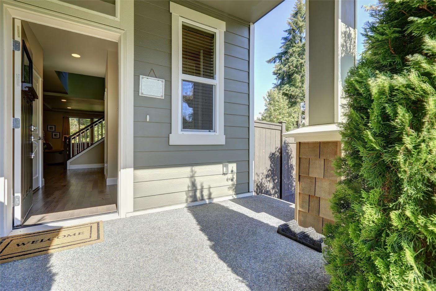 Gray porch with a concrete floor, leading to an open doorway. There's a window on the wall, and green bushes on the right side.