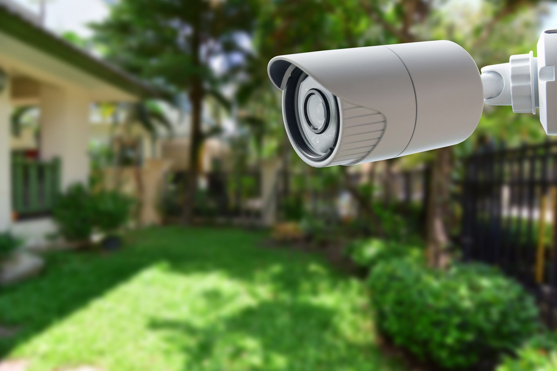 Security camera mounted outside, overlooking a green lawn and a house. Camera is light gray.