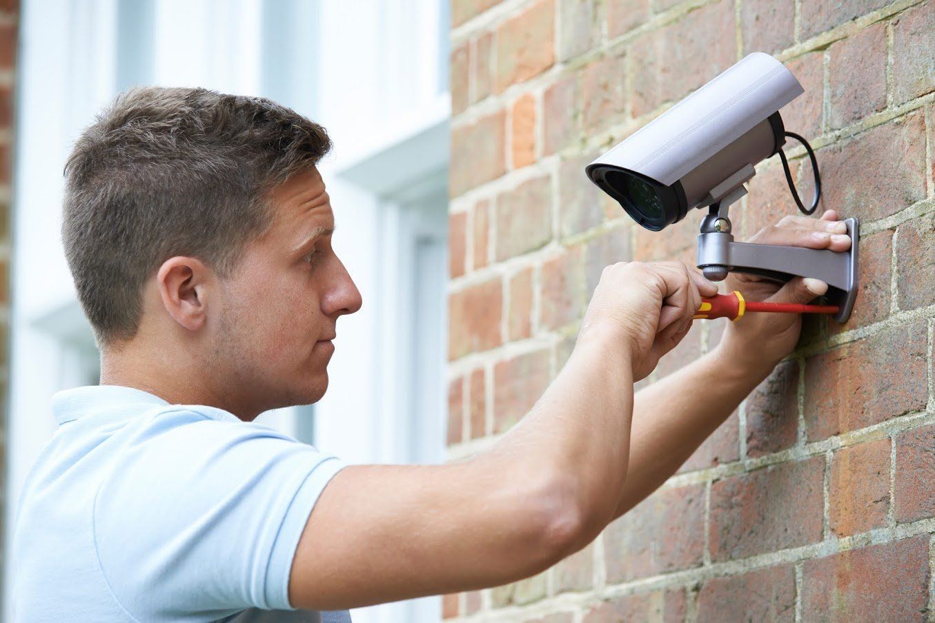 Man in light blue shirt installs a security camera on a brick wall with a screwdriver.