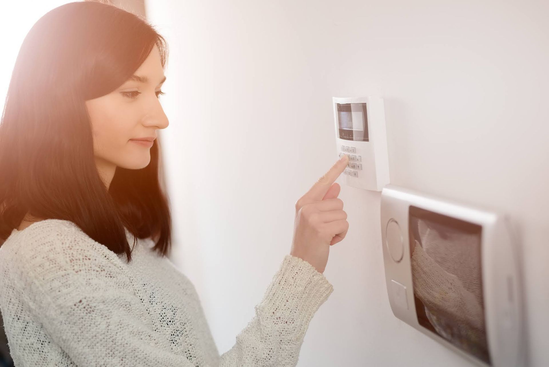 Woman in a sweater adjusting a white thermostat on a white wall, near a lower screen display; natural light.