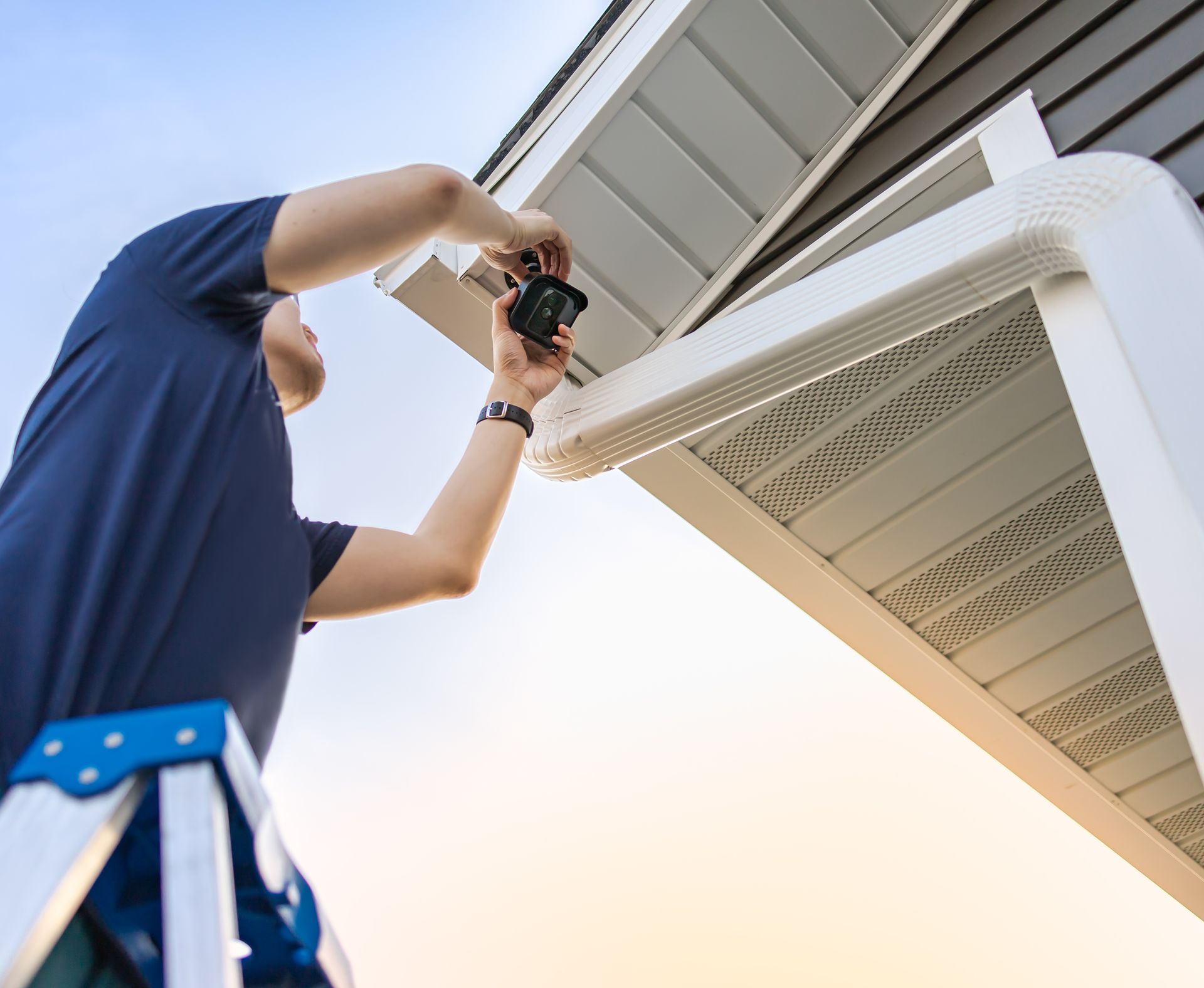 A technician on a ladder installing and adjusting a wireless security video camera underneath a roof