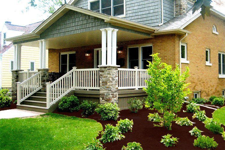 A two-story home with a porch, featuring brick siding and landscaping. Brown mulch surrounds green shrubs and lawn.