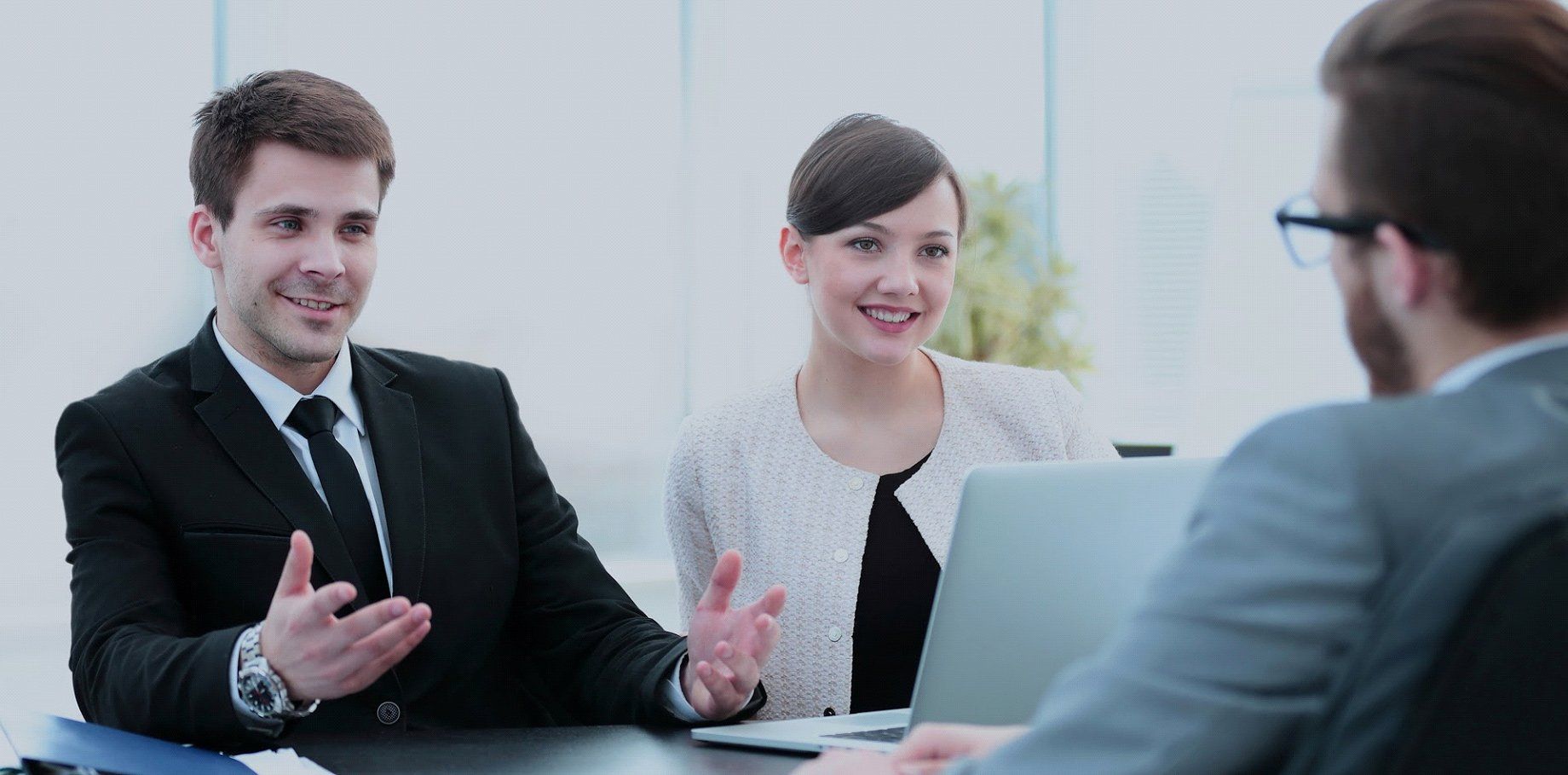 Three people in business attire are having a meeting in an office. The man on the left gestures with his hands, and the woman smiles.