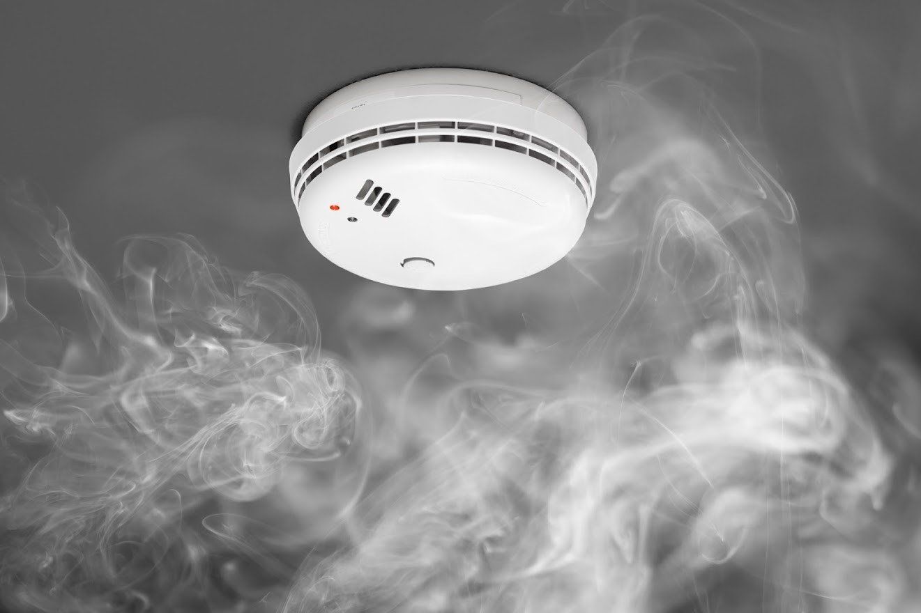Smoke detector on a ceiling, surrounded by grey smoke, with a red light illuminated.