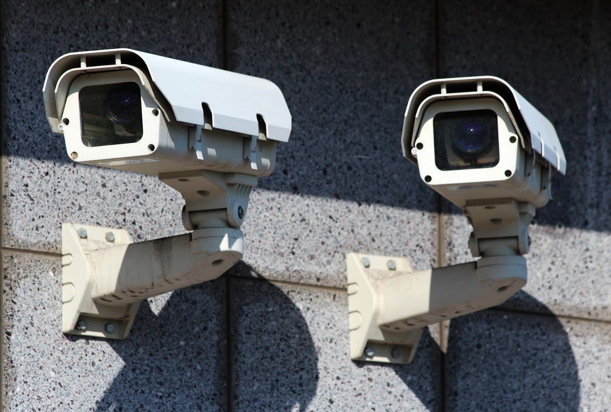 Two weathered, white security cameras mounted on a gray brick wall, angled outward.