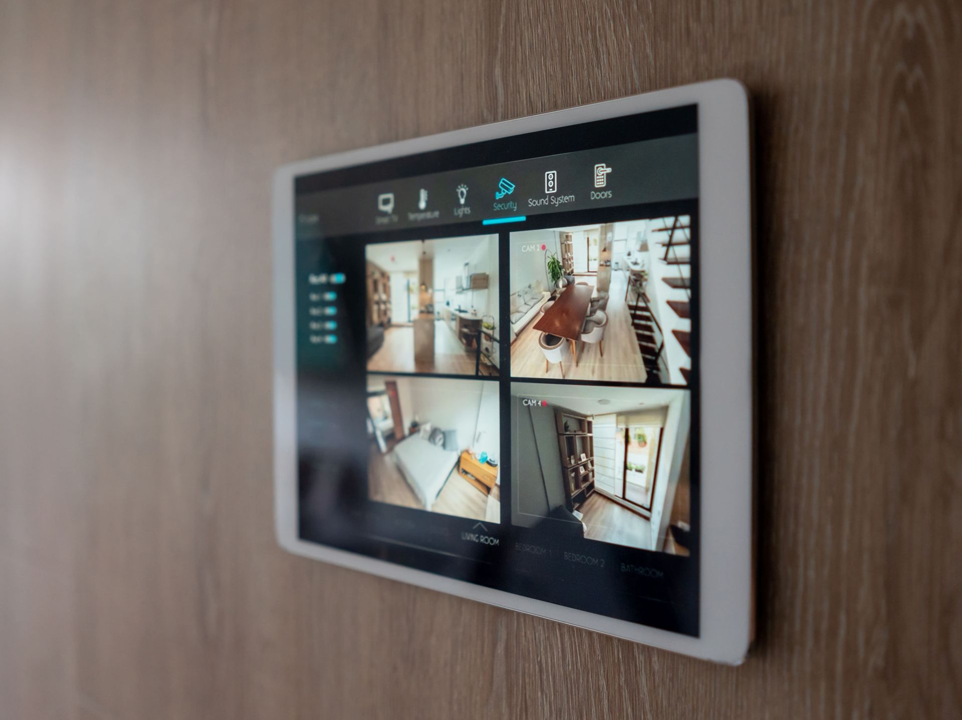 A white tablet mounted on a wooden wall displays a live feed from four security cameras within a home.