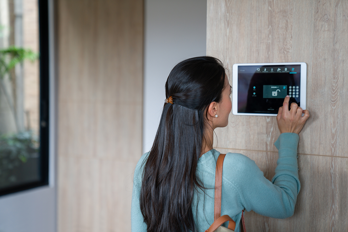 Woman interacting with a wall-mounted smart home control panel, inside a house with wooden paneling. She has long dark hair and wears a teal shirt.