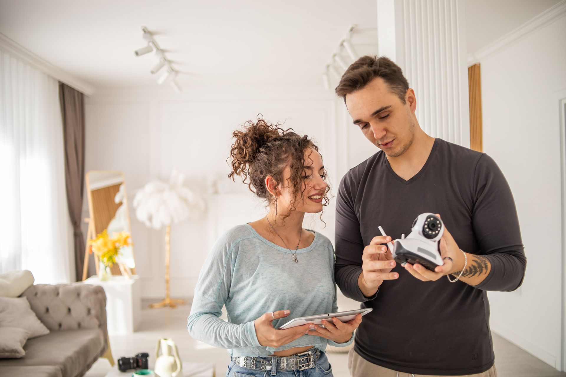 A security camera is hanging from the ceiling of a restaurant. A man and woman looking at a camera indoors. The man points at the camera while the woman holds a phone, both smiling.
