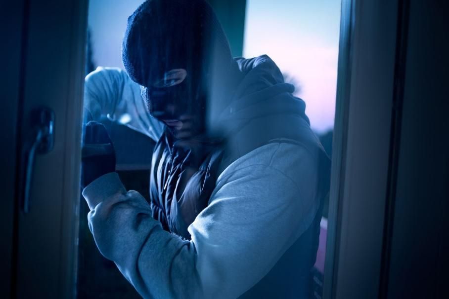 Person wearing a balaclava and hoodie breaking into a window at night, illuminated by dim blue light.