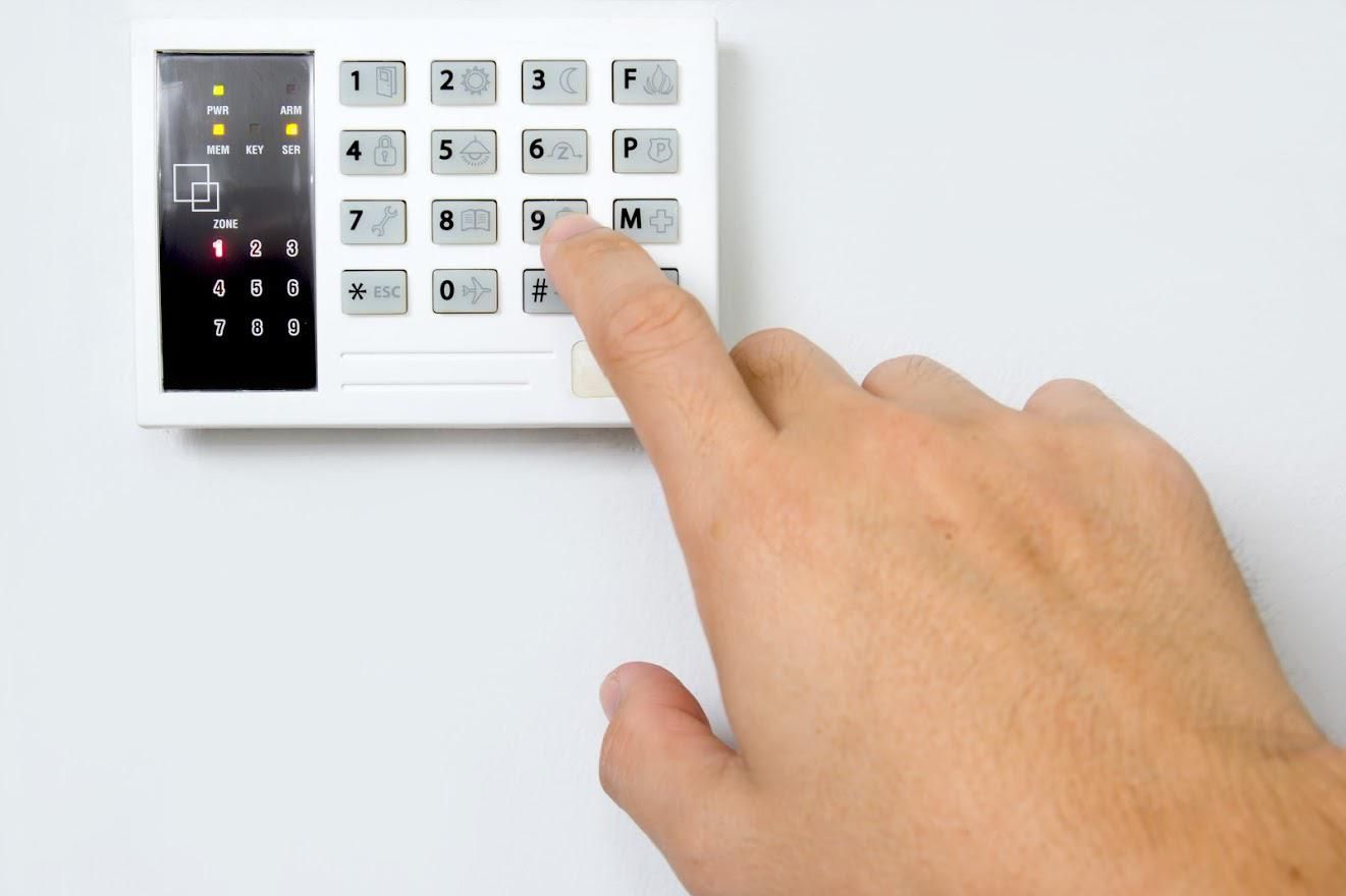 A hand entering a code on a white security system keypad mounted on a white wall.
