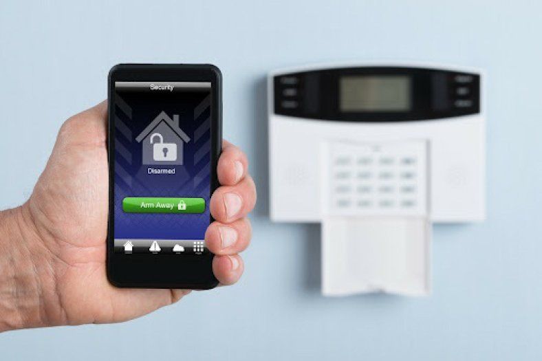 Hand holding a smartphone unlocking a house icon on the screen, next to a white security system panel on a light blue wall.