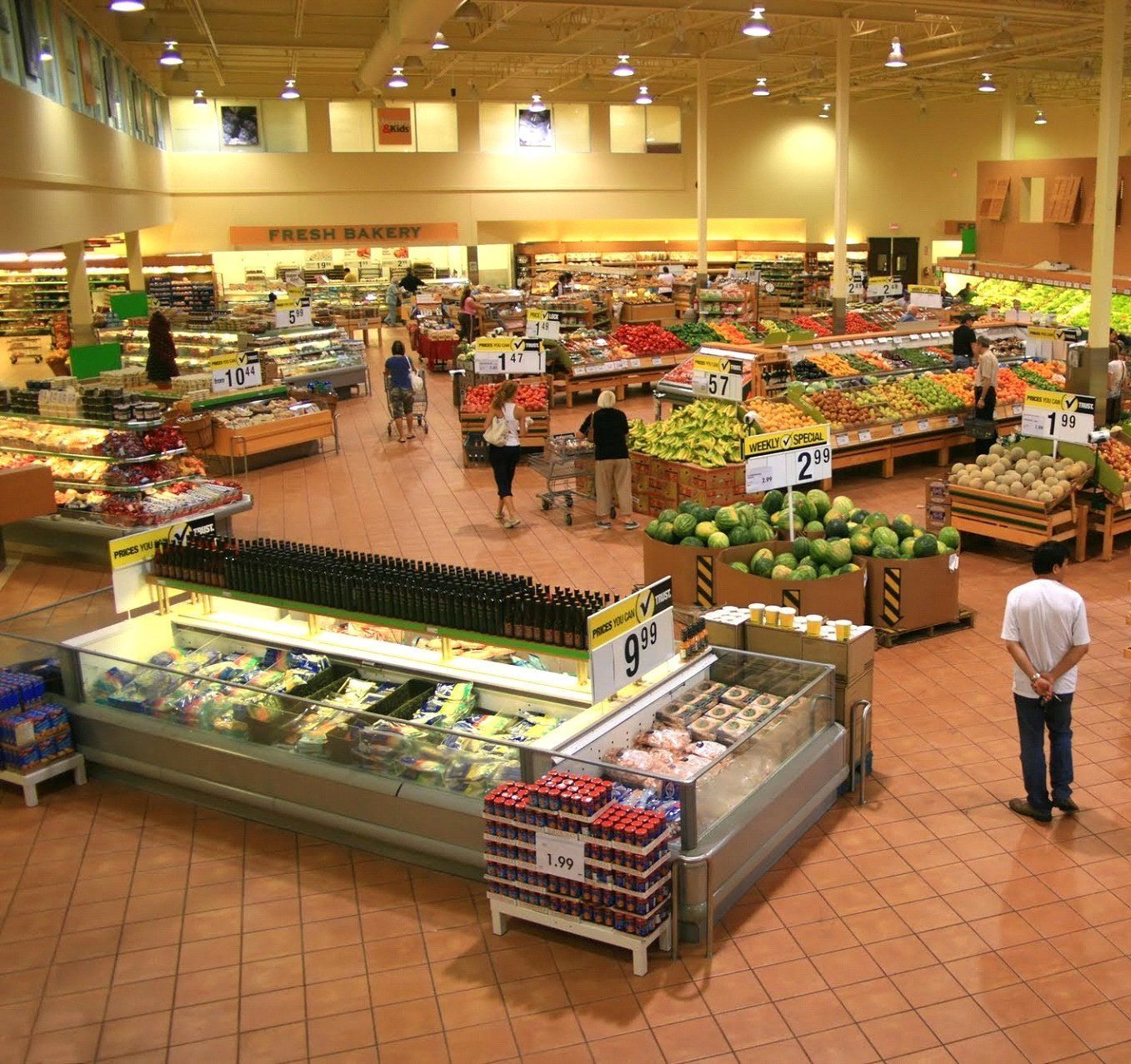 A wide-angle shot of a grocery store interior, showcasing produce, prepared foods, and customers shopping in brightly lit aisles.