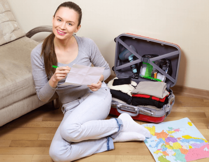 A woman packing a suitcase, making a list on a notepad while smiling. She's in a room with a sofa and a map.