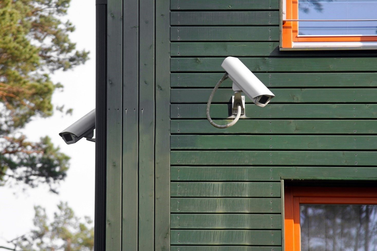 Two security cameras mounted on a green wooden building exterior. One camera is on the corner and the other is on the left side.