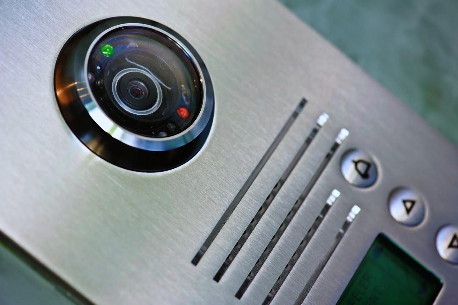 Close-up of a brushed metal intercom system with a camera, microphone slots, buttons, and a small green screen. The camera has a green and red LED.