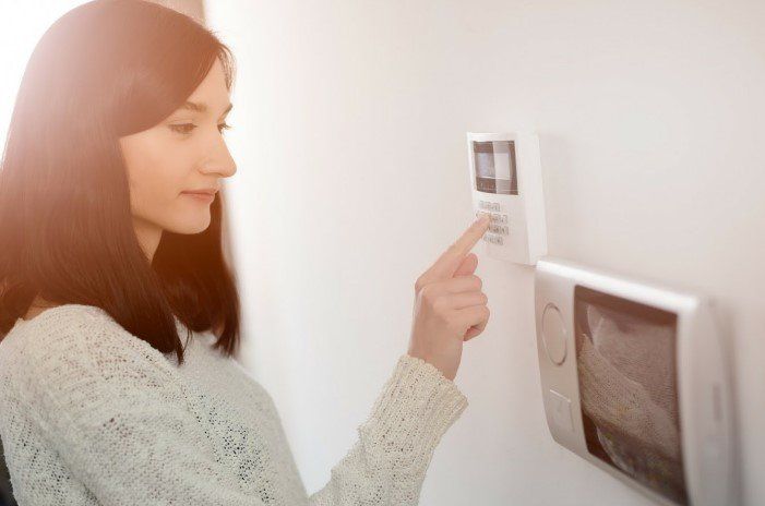Woman in a sweater pressing a button on a white wall-mounted thermostat. Another panel is below the thermostat.