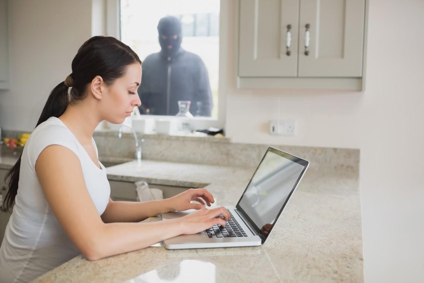 Woman using a laptop in a kitchen, unaware of a masked figure peering through the window.