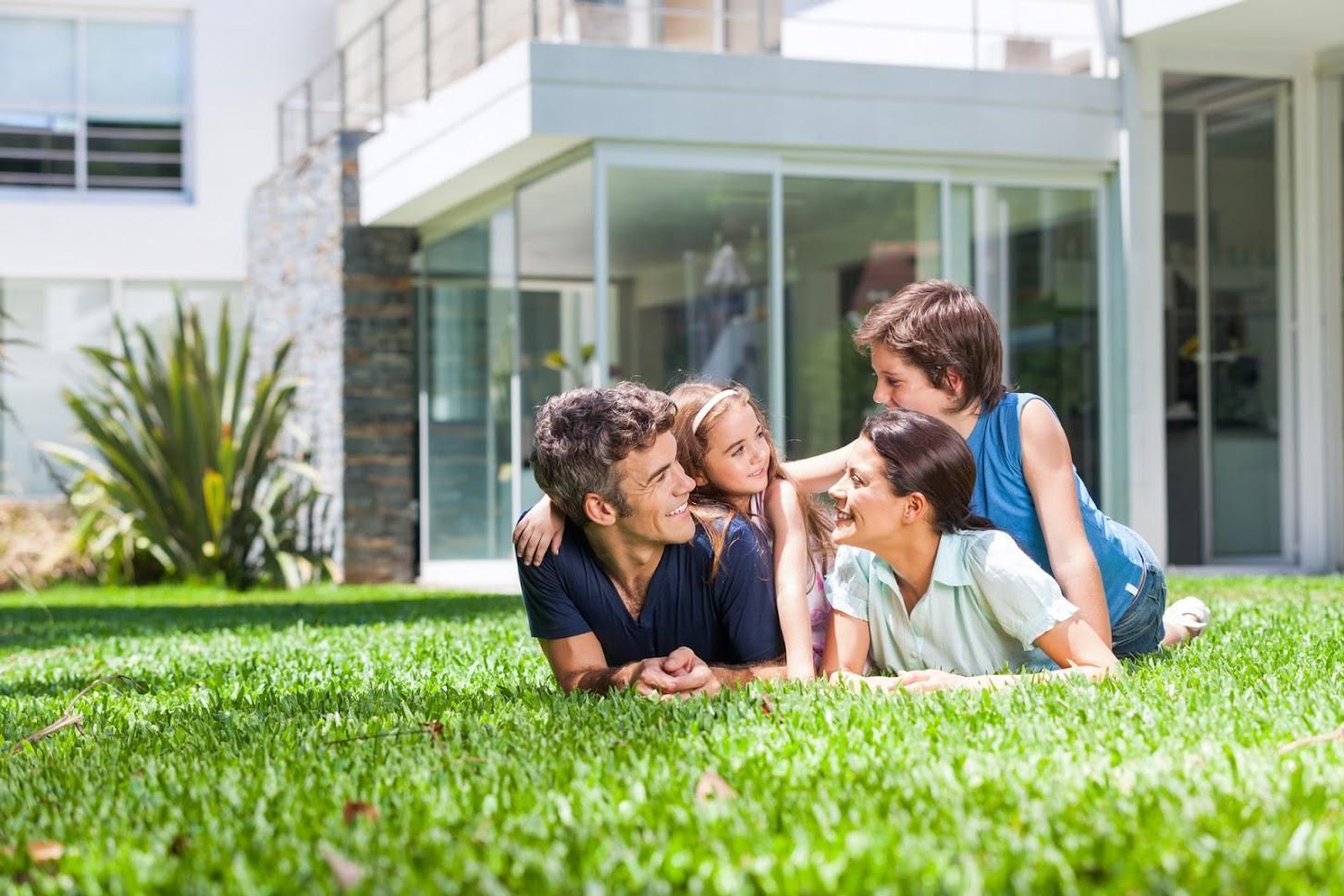 Family of four laying on grass in front of a modern house, smiling and looking at each other.
